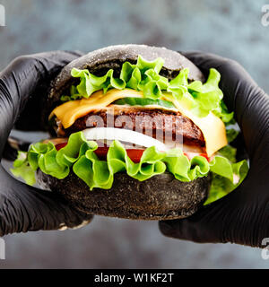 Les mains dans les gants noirs tenir un grand black avec burger de boeuf, fromage galette de marbre et des légumes frais. Vue rapprochée. Banque D'Images