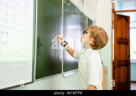 Petit garçon avec de grandes lunettes noires et une chemise blanche, debout près de l'école tableau noir avec un morceau de craie faisant face à une réflexion intelligente. Banque D'Images