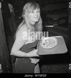 Années 1970, historiques, une femme blonde serveuse ou hôtesse avec un plateau de canapés lors du lancement d'un nouveau night club, London, UK. Banque D'Images