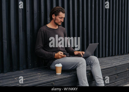 Je le photographe pigiste de l'emplacement ville avec thé ou café à l'écoute de la musique sur le casque, et sac travaille sur un ordinateur portable Banque D'Images