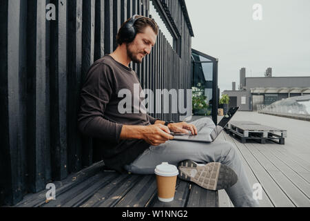 Je le photographe pigiste de l'emplacement ville avec thé ou café à l'écoute de la musique sur le casque, et sac travaille sur un ordinateur portable Banque D'Images