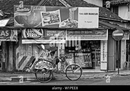 Yogyakarta, Indonésie - 2009.04.27 : un becak pilote dans son véhicule en attente de passagers en face d'une boutique Banque D'Images