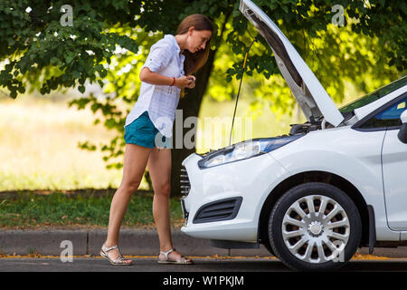 Jeune femme avec le smartphone à la recherche sur le moteur de voiture cassée Banque D'Images