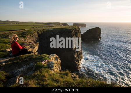 Une femme blonde dans une veste rouge et des lunettes de soleil se trouve sur le bord des roches pour le coucher du soleil sur les falaises de Kilkee et l'océan Atlantique, Kilkee, Banque D'Images