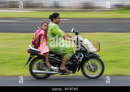Une femme dans une robe verte conduit un motorscooter avec une fille aux pieds nus dans une robe rayée rose et blanc et un sac à dos, assis derrière elle, Fongafale Isl Banque D'Images