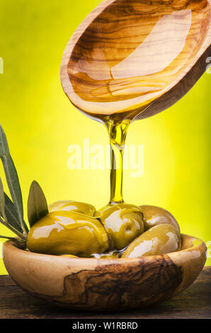 L'huile d'olive coulant dans un bol en bois d'olivier plein d'olives vertes sur fond vert. Banque D'Images