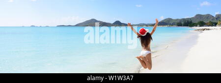 Woman with arms outstretched jumping at beach. Vue arrière de femelle est wearing Santa hat et bikini. L'insouciante est bénéficiant de Noël locations aux beaux jours. Banque D'Images