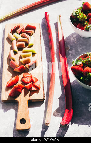 La rhubarbe et fraises fraîches coupées en morceaux sur un cutboard en bois sur une base en béton, des coups de table forte lumière directe avec de riches sh Banque D'Images