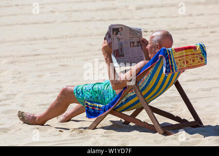 Homme assis dans une chaise longue à lire le journal sur la plage de Bournemouth, Dorset UK en Juillet Banque D'Images