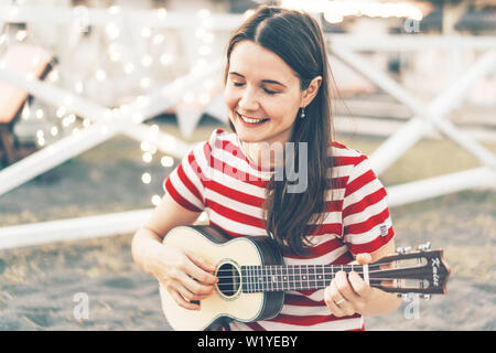 Une belle jeune brunette joue un ukulele guitare acoustique, un instrument de musique hawaïenne traditionnelle, festival de musique en plein air d'été, heureux au chaud même Banque D'Images