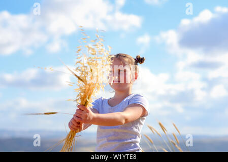 Happy little girl walking in golden wheat, tenant des pics de blé et les oreilles de l'avoine. La beauté de la nature, le ciel bleu, les nuages blancs et champ de blé. Banque D'Images