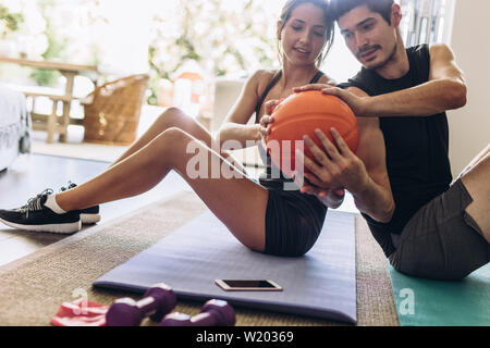 Jeune couple en bonne santé faisant des exercices avec un ballon à la maison. Couple assis dos à dos sur un tapis de yoga exerçant en passant la balle à l'autre. Banque D'Images