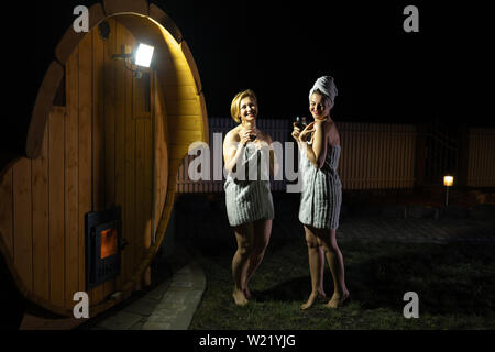 Deux copines aux femmes qui boivent du vin rouge à partir de verres outsite près d'un sauna mobile en bois avec juste les serviettes Banque D'Images