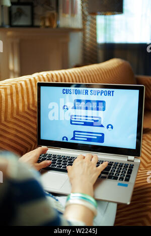 Femme au foyer assis sur le divan dans la maison moderne en chaude journée d'été ensoleillée en utilisant ai service client sur un ordinateur portable. Banque D'Images