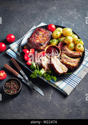 Côtes de porc grillées recouvertes de sauce barbecue servi avec des tomates et de l'eau chaude les pommes de terre servies sur un plateau noir sur une table, vue verticale Banque D'Images