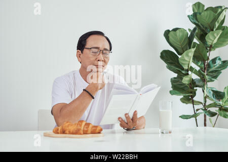 Vieil homme boire du lait et de la lecture livre tout en ayant le petit déjeuner Banque D'Images