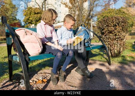 Petit garçon et fille écoliers lecture livre, assis sur un banc, des enfants avec des sacs à dos, journée d'automne ensoleillée Banque D'Images