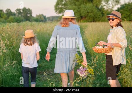 Femme avec deux filles marcher dans un pré avec un panier d'œufs, vue de dos, heure d'or. Banque D'Images