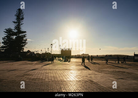 Coucher du soleil dans une place urbaine à Essaouira, Maroc, une ville portuaire et station balnéaire sur l'océan Atlantique avec les gens marcher silhouetté contre le soleil Banque D'Images