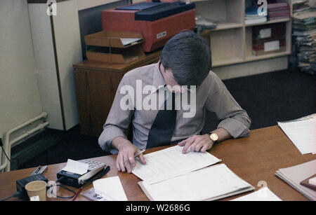 Années 1970, historique, un homme, fin des années 20, ou début des années 30 dans l'âge dans un bureau, assis à un bureau de travail, un directeur financier, ou peut-être un comptable comme il est à l'aide d'un petit calculateur électronique pour ajouter les chiffres des grands livres ou journaux imprimés, England, UK. Banque D'Images