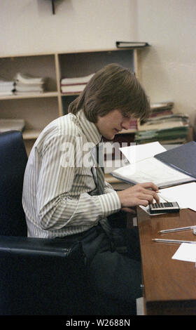 Années 1970, historique, un homme, fin des années 20, ou début des années 30 dans l'âge dans un bureau de travail, assis à un bureau, un directeur financier ou un comptable peut-être qu'il est à l'aide de calculatrice pour ajouter des chiffres, England, UK. Banque D'Images