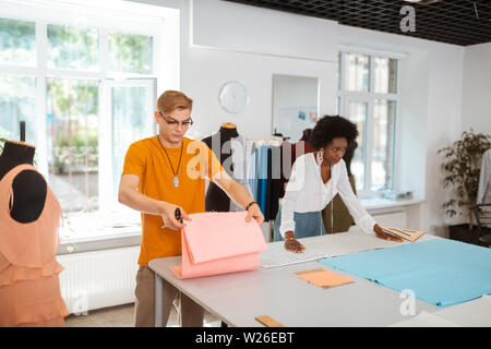 Travailler avec le tissu. Les jeunes d'adapter la tenue des ciseaux et de tissu dans ses mains tout en se tenant au bureau à côté de son collègue Banque D'Images