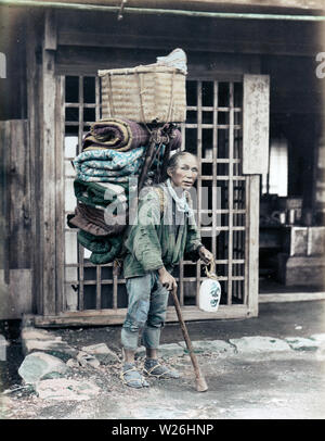 [ 1890 - Japon ] - Transport des bagages Porter Porter un transport des bagages, des courtepointes, futon, semble-t-il, sur un cadre en bois sur le dos. Il tient une canne dans sa main droite et un tokkuri (alcool) dans sa main gauche. Il y a une petite serviette autour du cou. Dans l'arrière l'avant d'une maison ou d'un atelier peut être vu. 19e siècle vintage albumen photo. Banque D'Images