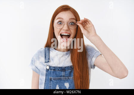 Rousse animé impressionné happy girl porter des salopettes en denim smiling étonné cheerful touch lunettes caméra regarder étonné et heureux, exprimer ma stupéfaction un Banque D'Images