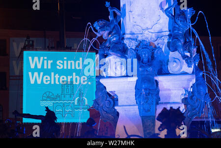 Augsburg, Allemagne. Le 06 juillet, 2019. La fontaine d'Auguste est éclairé et sur la façade du bâtiment de l'administration de la ville le lettrage "Wir sind Welterbe' est prévu. Augsbourg a été inclus dans la Liste du patrimoine mondial de l'UNESCO en raison de son système d'eau historiques. Credit : Stefan Udry/dpa/Alamy Live News Banque D'Images