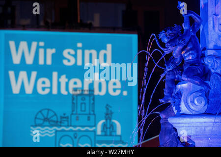 Augsburg, Allemagne. Le 06 juillet, 2019. La fontaine d'Auguste est éclairé et sur la façade du bâtiment de l'administration de la ville le lettrage "Wir sind Welterbe' est prévu. Augsbourg a été inclus dans la Liste du patrimoine mondial de l'UNESCO en raison de son système d'eau historiques. Credit : Stefan Udry/dpa/Alamy Live News Banque D'Images