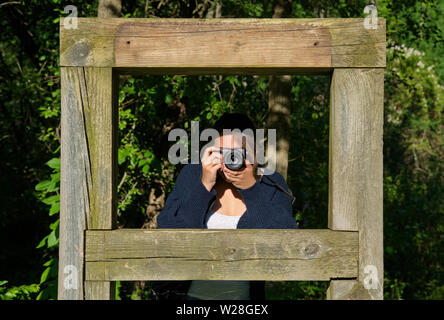 Middletown, CT USA. Jun 2019. Yong woman pointing la nouvelle caméra type mirrorless à partir d'un cadre en bois dans les bois. Rédaction d'illustration Banque D'Images