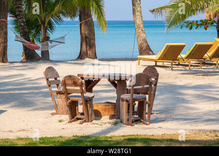 La plage de sable tropicale avec table et chaises en bois, chaises longues, hamac et le vert des palmiers sur fond de mer bleue, la Thaïlande. Billet d Banque D'Images