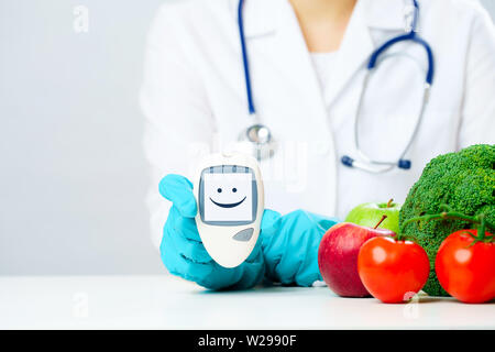 Photo de médecin en blouse et des gants en caoutchouc avec diabete montrant smiley à table avec des légumes sur fond gris Banque D'Images