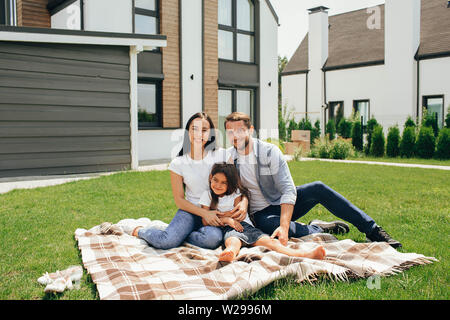 Heteroseksual happy family sitting on grass près de leur nouvelle maison. Famille assis sur la pelouse, l'achat de nouveaux big home . Banque D'Images