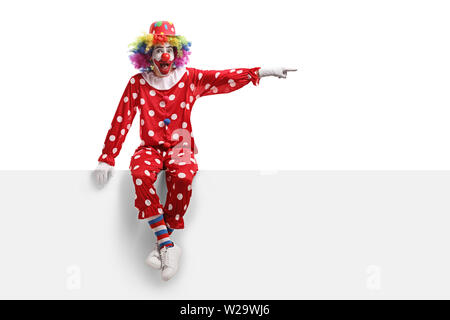 Longueur totale d'un clown assis sur un panneau blanc et pointant vers le côté isolé sur fond blanc Banque D'Images