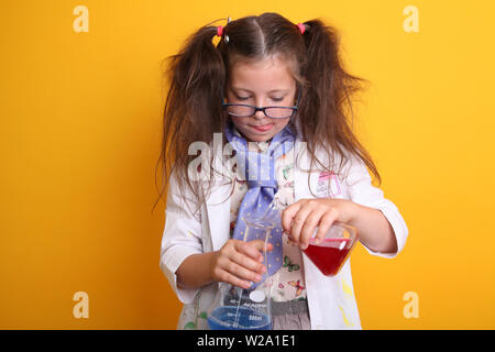 M. - Science Geek jeune fille en lunettes âge 7 mesure des produits chimiques liquides dans des béchers scientifiques borosilicatés sur fond jaune espace de copie en arrière-plan Banque D'Images