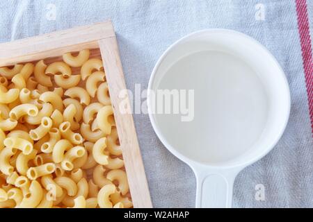 La nourriture et la cuisine, vue de dessus et non cuits ou secs pâtes macaronis coudés Gomiti au plateau en bois avec une tasse à mesurer en plastique. Banque D'Images