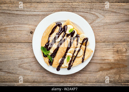 Crêpes ou blinis avec des tranches de banane et sauce au chocolat sur plaque blanche, des planches en arrière-plan. Vue d'en haut Banque D'Images
