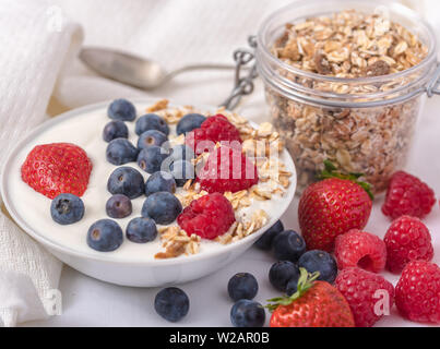 Le yogourt dans un bol blanc avec le gruau et les fraises, bleuets et framboises sur le dessus sur fond blanc. Banque D'Images