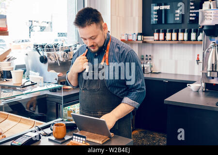 Jeune homme caucasien homme barbu en restaurant café barista caissier au travail. Vendeur sur le bloc de touches pour l'acceptation de paiement client client achat. Sm Banque D'Images