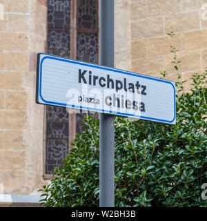 Lettrage enseignes bilingues Place de l'Église, en allemand, Kirchplatz, et italien piazzo chiesa. Mur de l'église en arrière-plan. Banque D'Images