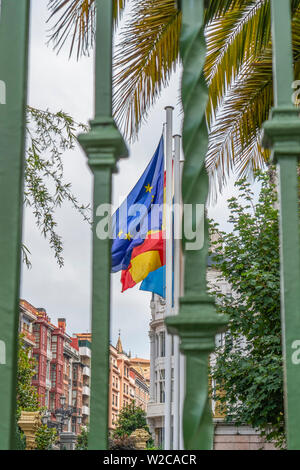 Drapeaux de l'Union européenne, l'Espagne, région des Asturies sur la façade d'un bâtiment ancien, derrière une grille de clôture verte. Contre les branches vertes de palm Banque D'Images