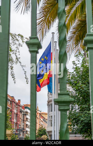 Drapeaux de l'Union européenne, l'Espagne, région des Asturies sur la façade d'un bâtiment ancien, derrière une grille de clôture verte. Contre les branches vertes de palm Banque D'Images