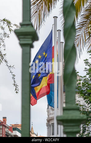 Drapeaux de l'Union européenne, l'Espagne, région des Asturies sur la façade d'un bâtiment ancien, derrière une grille de clôture verte. Contre les branches vertes de palm Banque D'Images