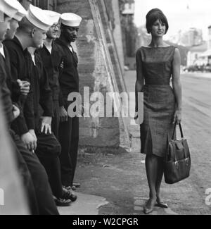 La mode dans les années 60. Un groupe de marins américains sont debout sur le quai en permission et fait un bel ajout à l'analyse de la mode. Une jeune modèle passe entre eux portant une robe et sac à main et chaussures. La Suède 1964 ref BV59-4