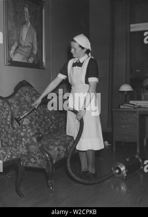 L'aspirateur dans les années 40. Une jeune servante est l'aspirateur à fond de la table. Elle est vêtue de sa tenue de travail blanche. Suède 1940 Banque D'Images