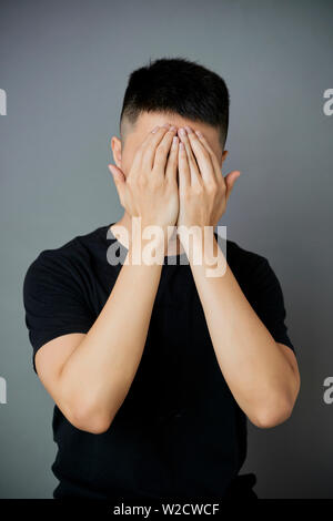 Portrait of young man standing in T-shirt noir et de cacher son visage avec ses mains isolé sur fond gris Banque D'Images