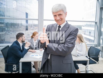Confiant mature businessman showing thumb up and smiling , équipe de ses collègues assis à la table dans l'arrière-plan Banque D'Images