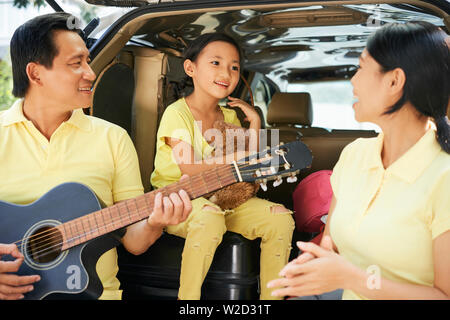 La famille asiatique debout près de sa voiture à l'extérieur et passer du temps ensemble drôle avec mère fille chantant une chanson alors qu'heureux père jouant gui Banque D'Images