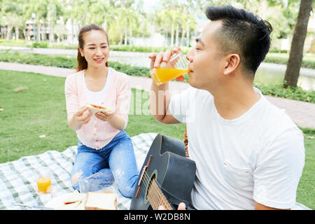 Young Asian man with guitar de boire du jus d'orange avec femme assise près de lui et maintenant ils sont en sandwich pique-nique dans le parc Banque D'Images